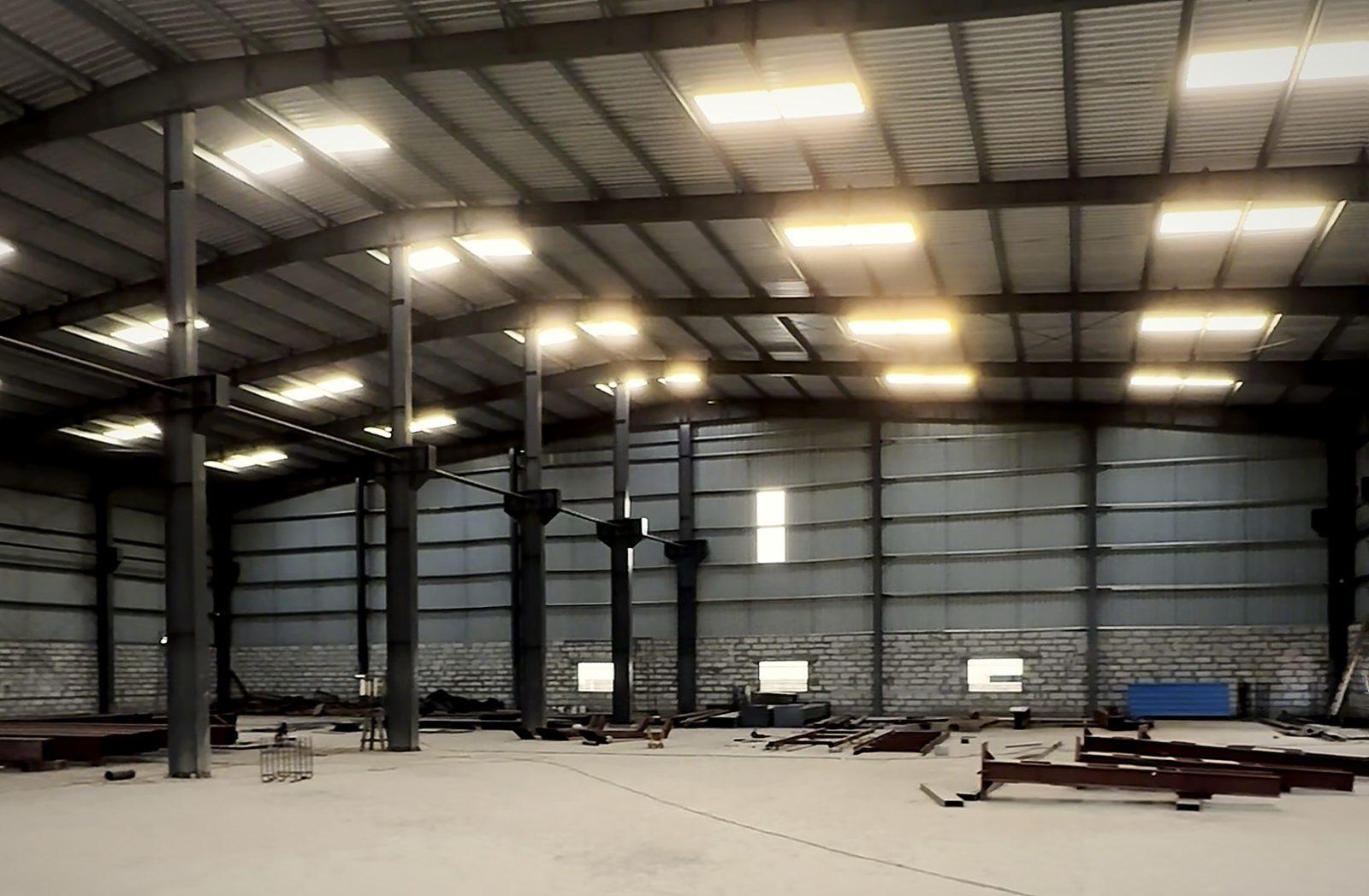 Inside the main fabrication bay at Kudumalakunte: a tall steel-framed shed with roof lights, with fabricated members laid out on the shop floor